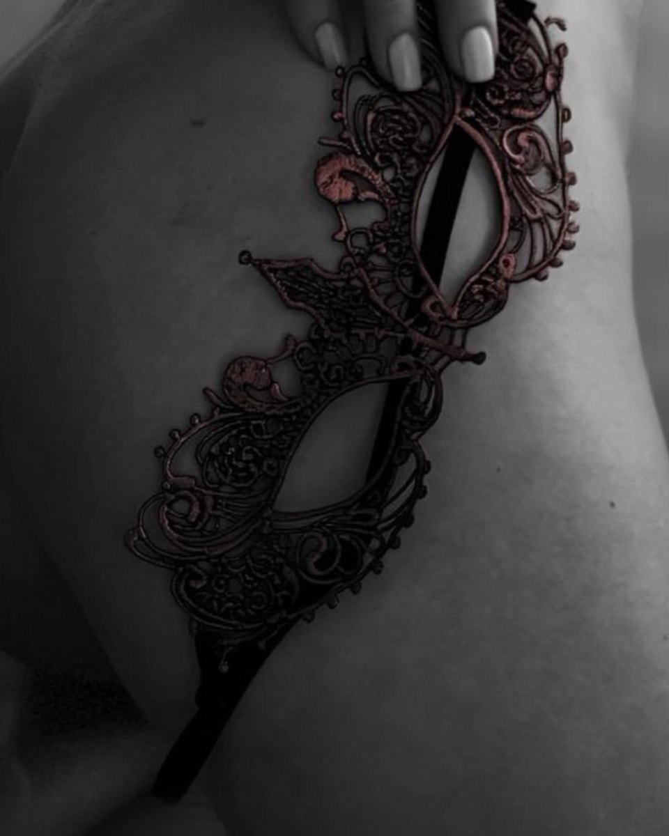 A hand holding a filigree masquerade mask against bare skin, in black and white